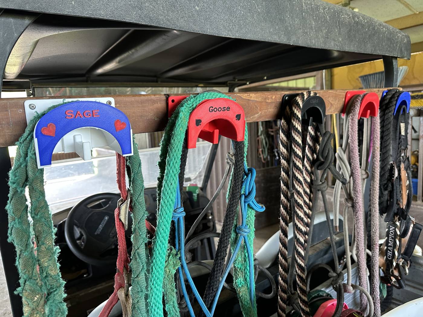 Custom 3D-printed halter and bridle holders mounted on a golf cart rail, each labeled with a horse's name